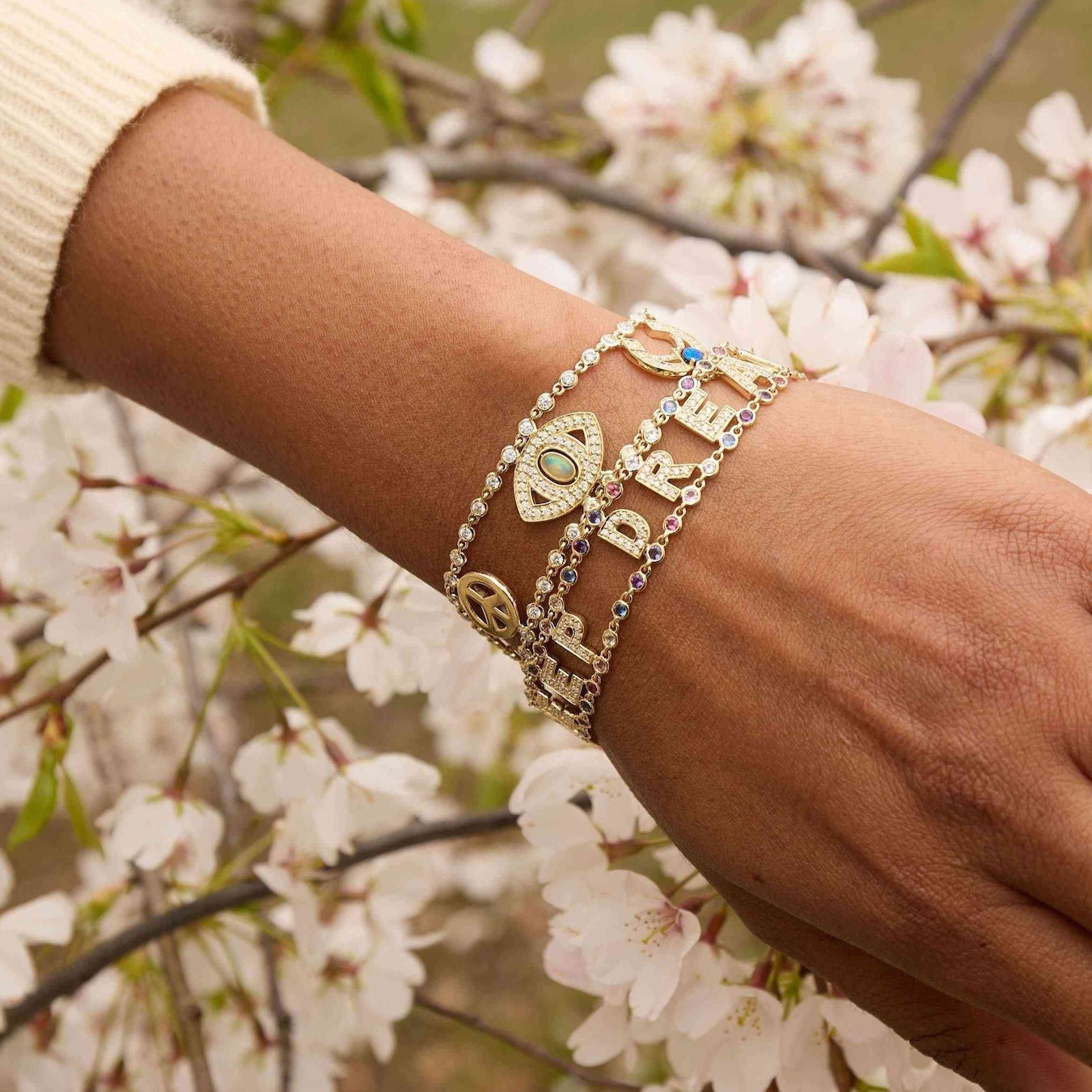 Hand wearing multiple gold bracelets with charms against a cherry blossom background