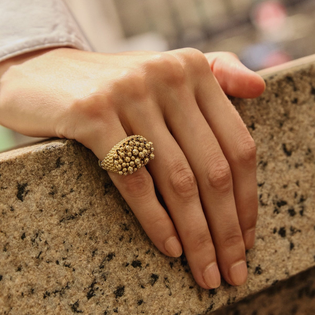 Hand wearing a gold ring on a stone ledge with a blurred urban background